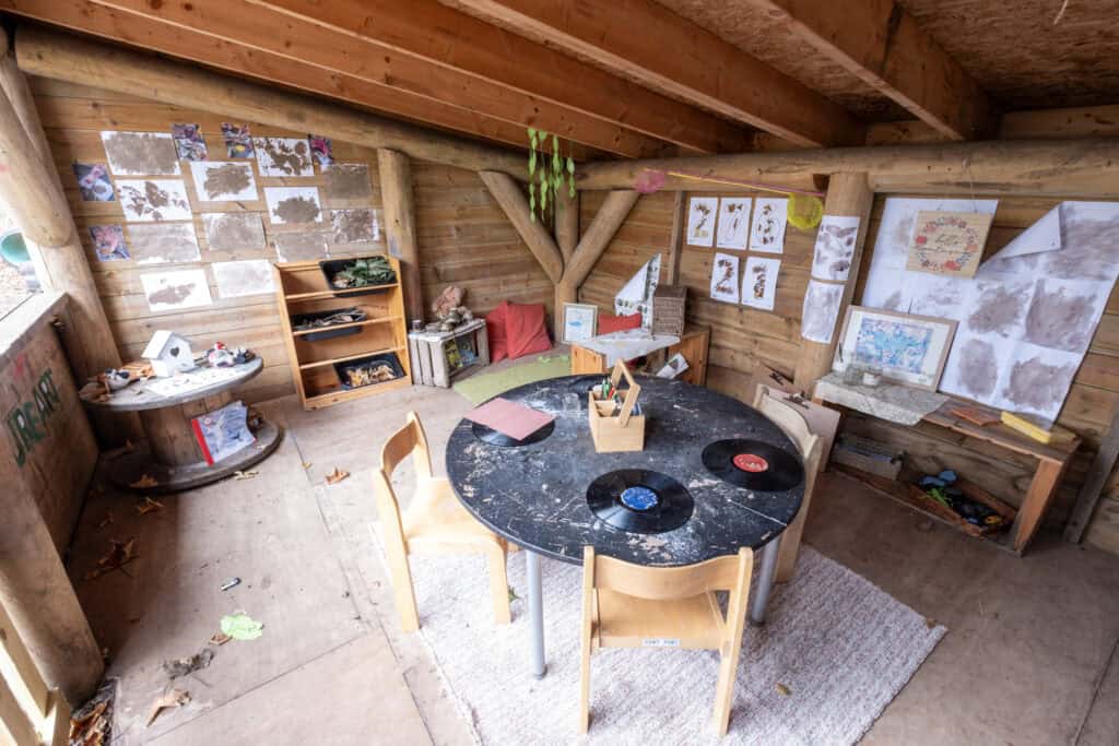 1. Well-loved rustic childcare art and activity nook in a wooden cabin, featuring tables, chairs, artworks, and educational supplies for early childhood development.