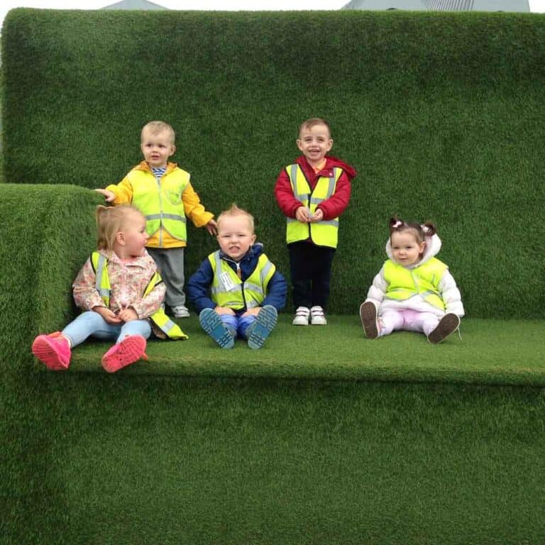 Playful young children sitting outdoors on a lush green artificial grass structure, wearing high-visibility safety vests, enjoying outdoor activities at Thrive Childcare.