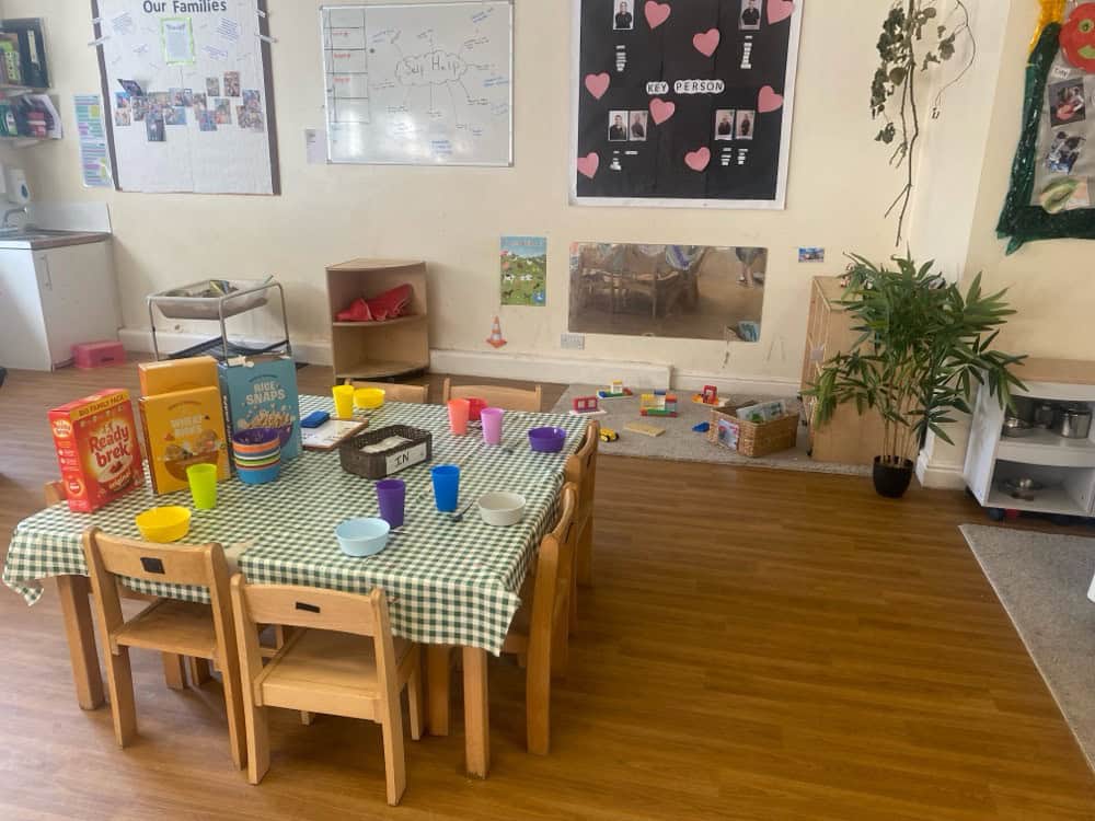 Brightly decorated nursery dining area with a checkered tablecloth, colourful cups and bowls, and children's artwork on the walls.