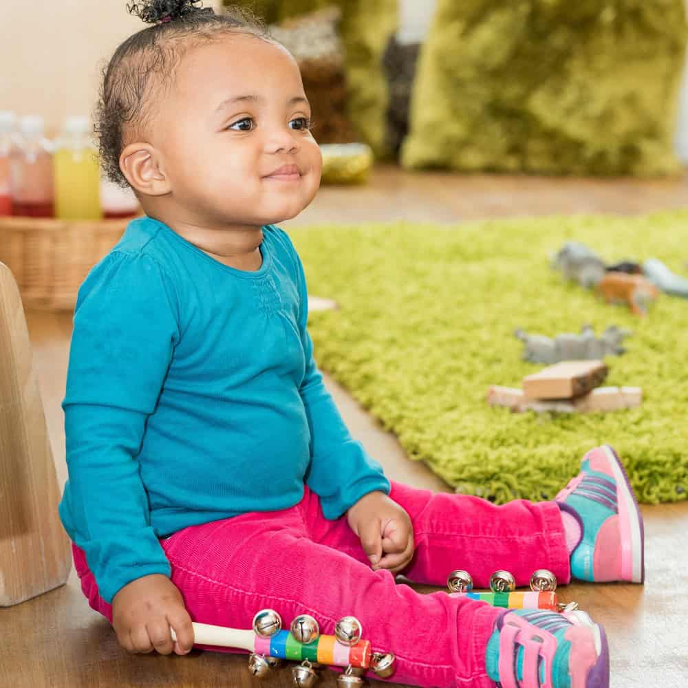 1. Adorable toddler girl sitting on wooden floor with musical jingle toys, bright infant care environment, joyful early childhood education, thriving childcare services, colourful educational toys.