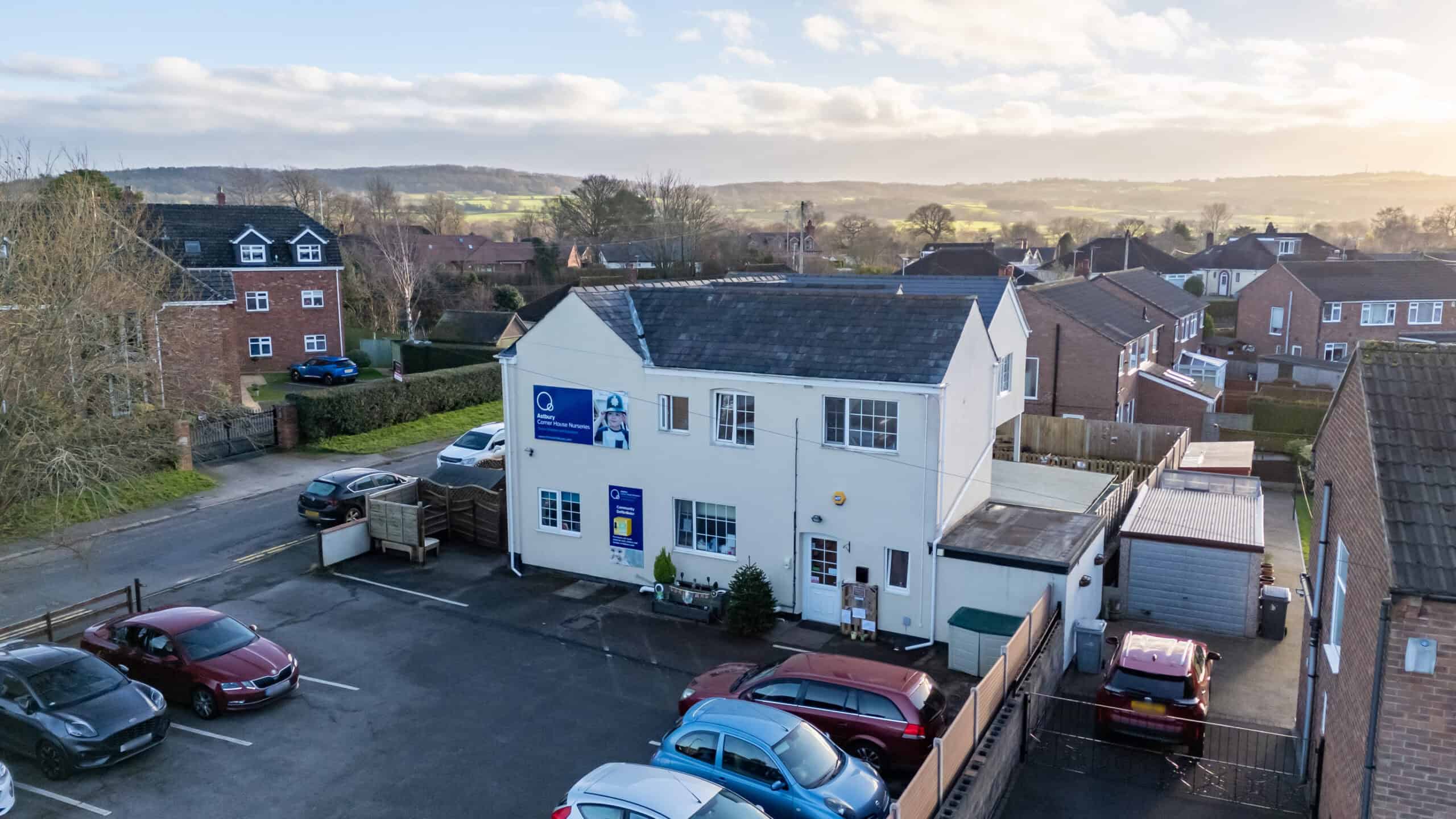 Aerial view of Corner House Astbury, a white childcare building with parking, situated in a residential area with houses and greenery in the background.