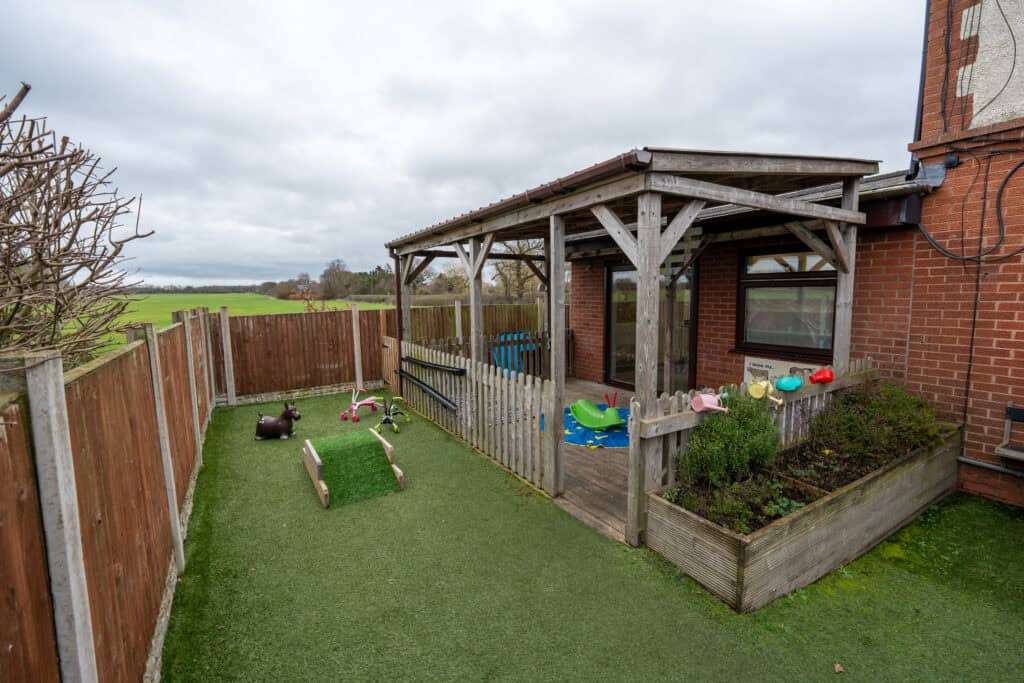 Bright outdoor play space at Thrive Grosvenor House with a wooden shelter, garden beds, and various toys for children’s outdoor activities.