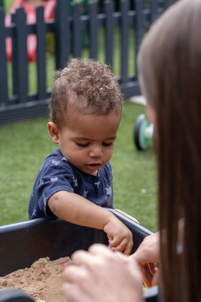 1. Young child playing in outdoor sandbox at Thrive Childcare, engaging in sensory and imaginative play, while supervised by a caregiver in a safe, nurturing environment.