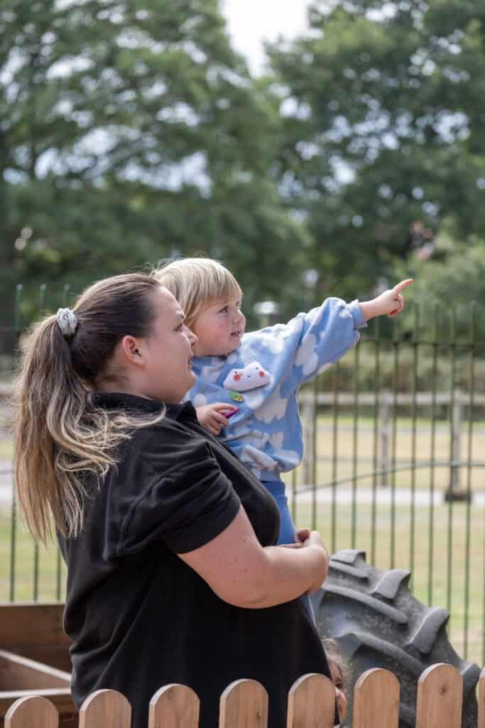 1. A young child with a caregiver outdoors at Thrive Childcare, engaging in play, surrounded by green trees and outdoor equipment, promoting early childhood development and outdoor learning.