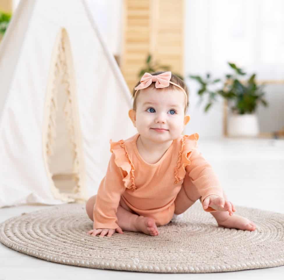 Bright-eyed baby girl in a soft peach outfit and pink headband crawling on a neutral rug in a modern, bright childcare setting with natural decor at Thrive Childcare.