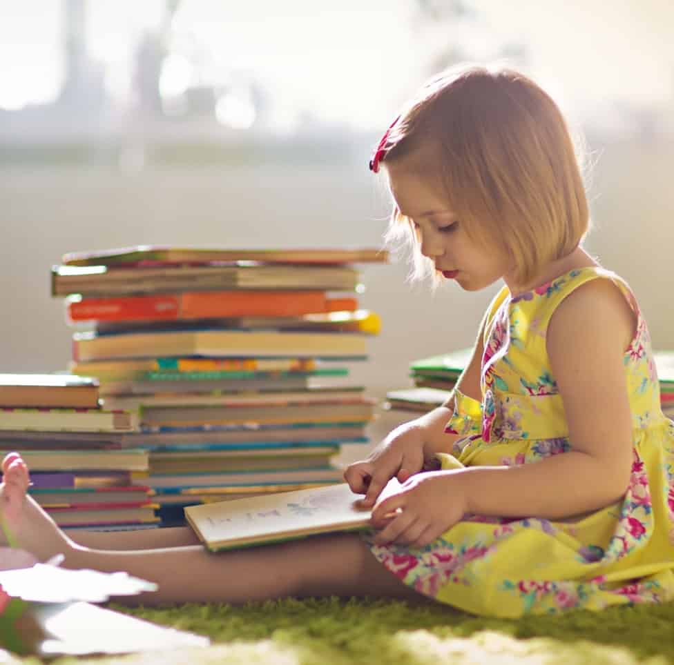 1. Young girl reading a children's book in a bright, safe childcare setting with stacks of colourful books, promoting early childhood education and nurturing environments at Thrive Childcare.