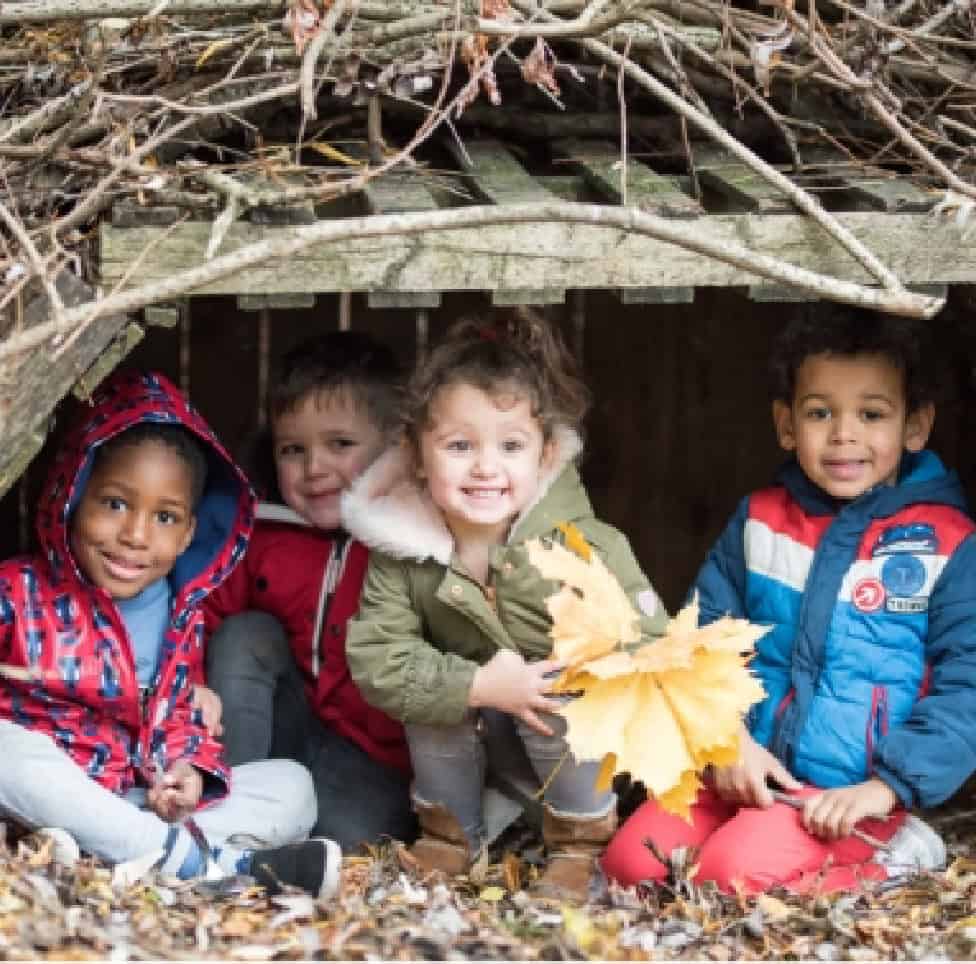 Playful children exploring outdoor nature play in a wooden den with autumn leaves, promoting early childhood development, outdoor learning, and creative childcare activities at Thrive Childcare.