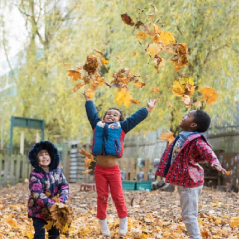 Playing children outdoors in autumn at Thrive Childcare, with leaves falling and children enjoying nature. Warm clothing and happy faces highlight a fun, safe childcare environment.
