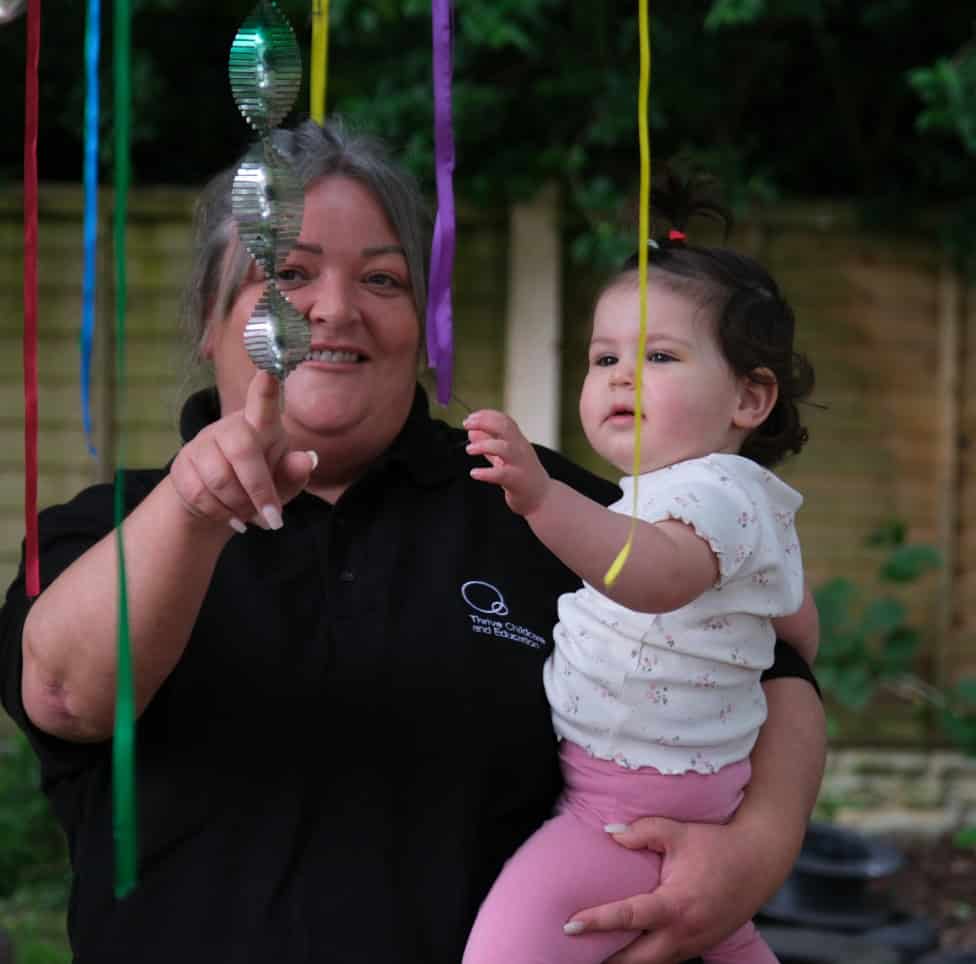 Brightly smiling childcare worker holding a young girl with colourful streamers at Thrive Childcare outdoor event, showcasing a nurturing environment for early childhood development and fun activities.