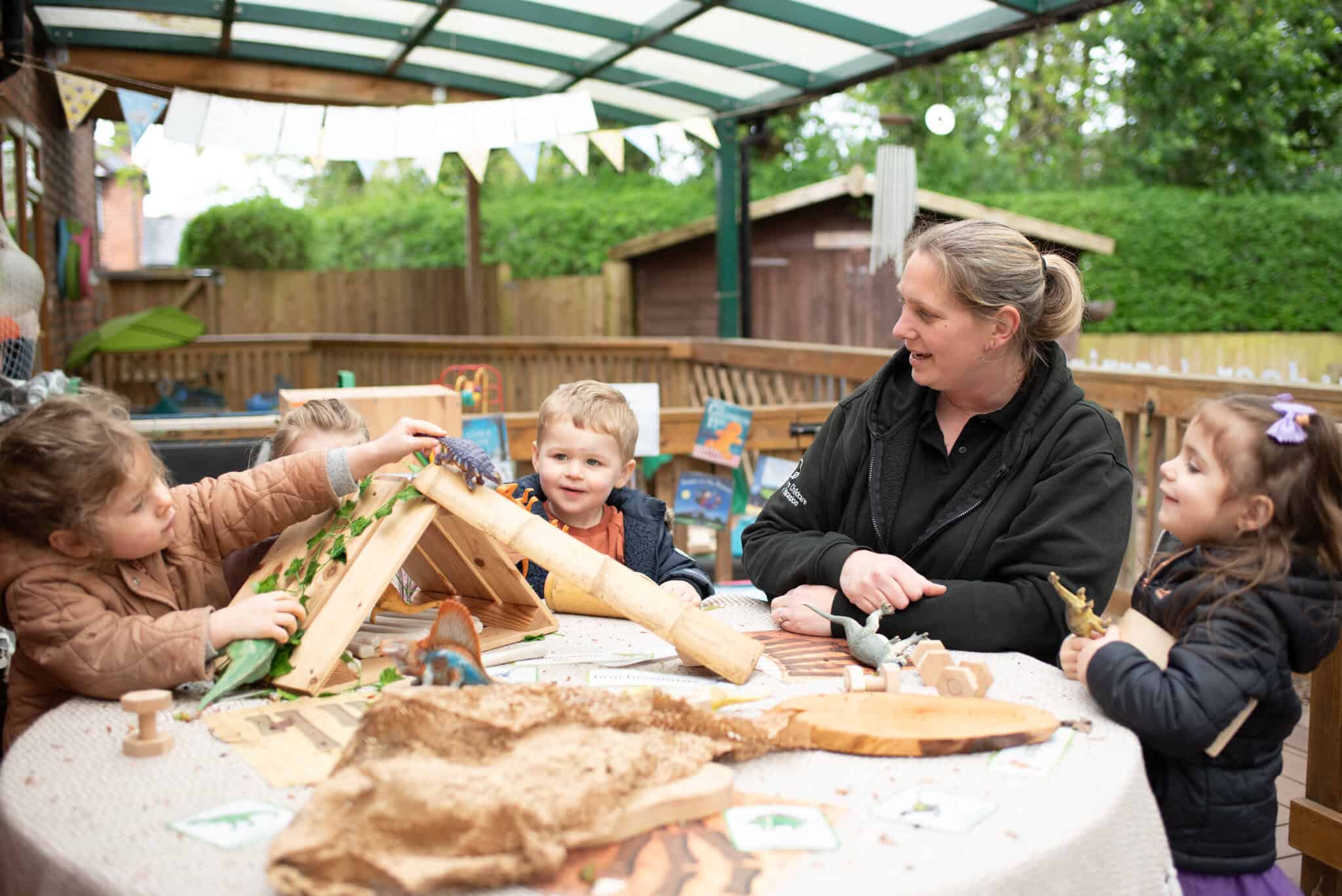 Bright cheerful outdoor play area at Thrive Childcare with children exploring nature-themed toys and educational activities under a shelter. A caring adult engages with children in a safe, nurturing environment.