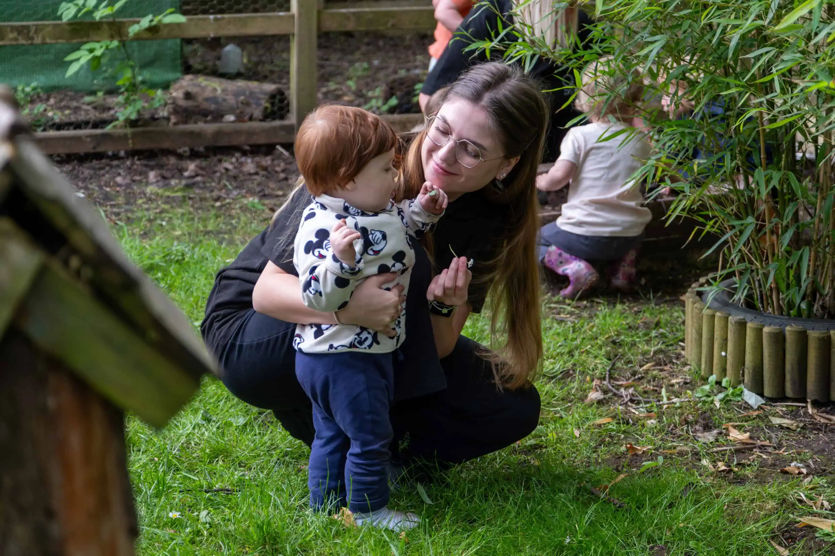 1. A caring childcare professional engaging with a young child outdoors in a garden setting at Thrive Childcare, promoting early childhood development and outdoor learning.