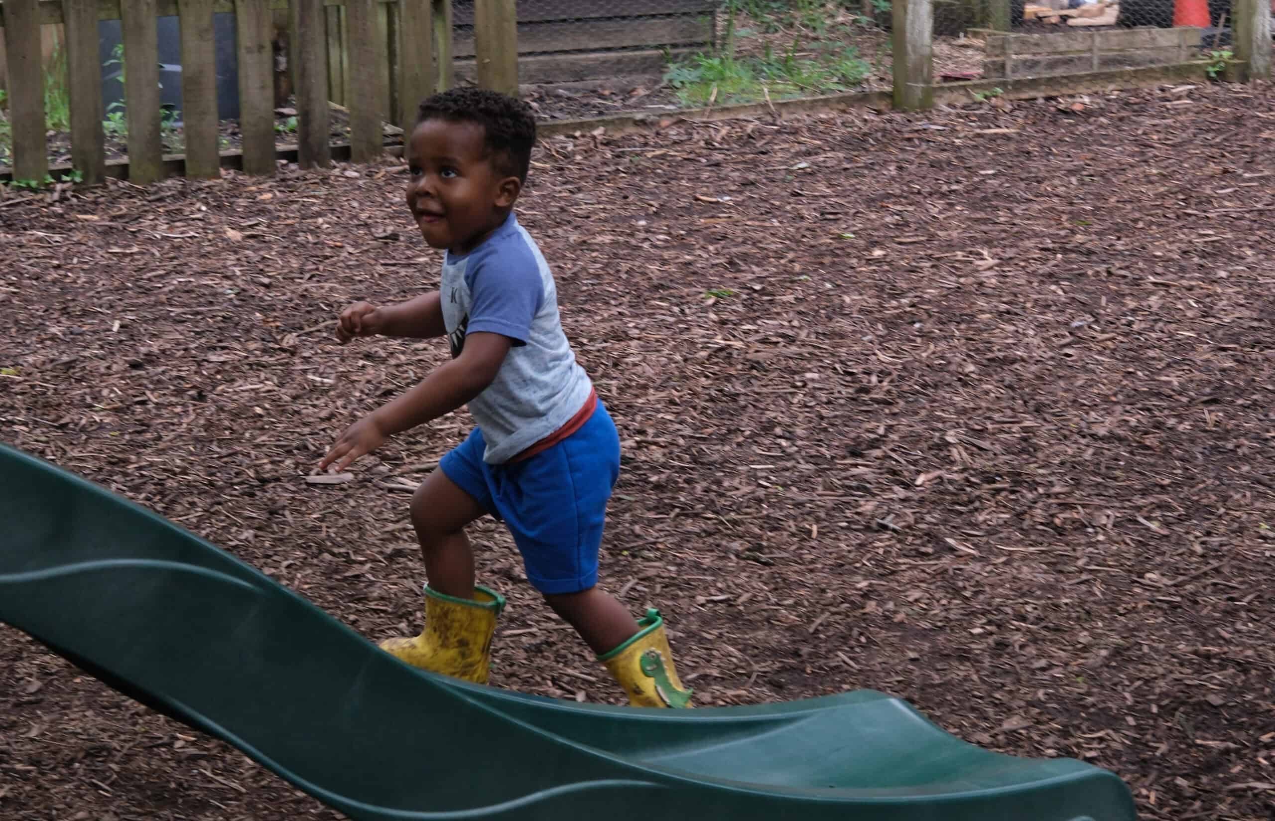 Young child playing outdoors at Thrive Childcare, running on a slide in a natural, wooded play area, promoting active and imaginative childhood play.