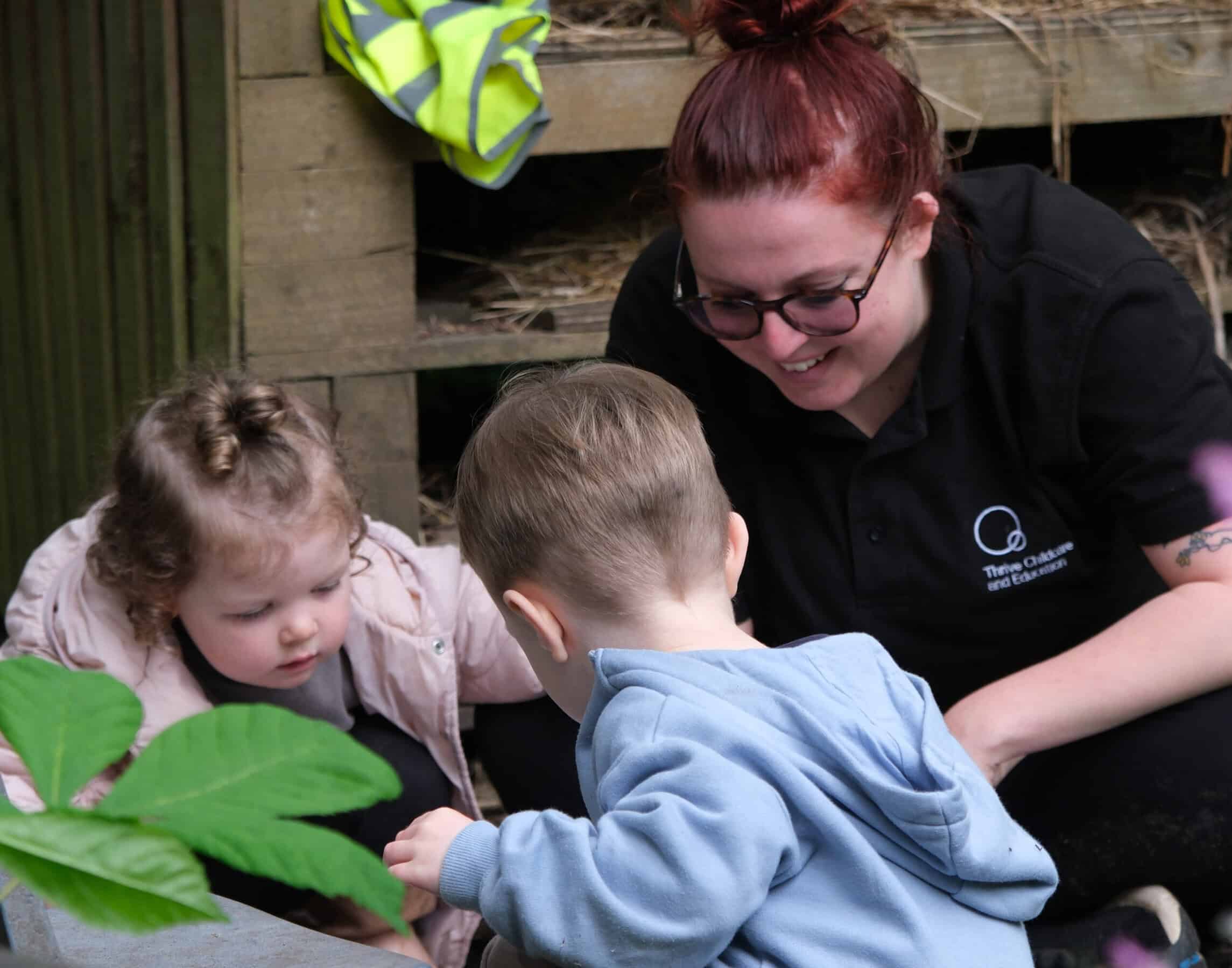 1. Friendly childcare worker engaging with young children outdoors in a natural play environment at Thrive Childcare.