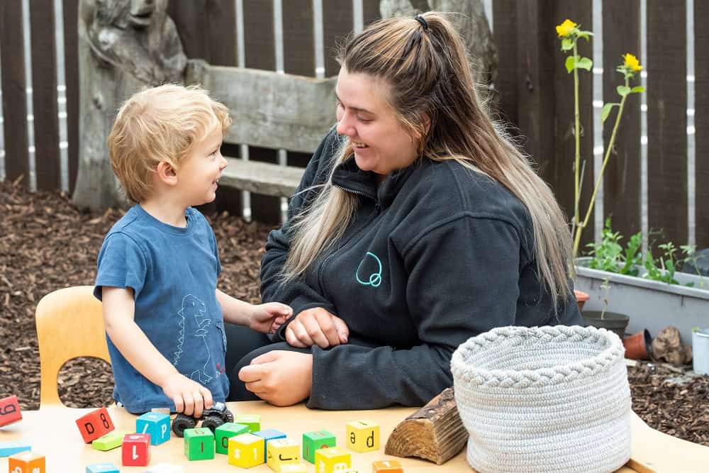Bright young child playing with colourful alphabet blocks outdoors with caring childcare professional at Thrive Childcare. Engaging learning environment for early childhood development.