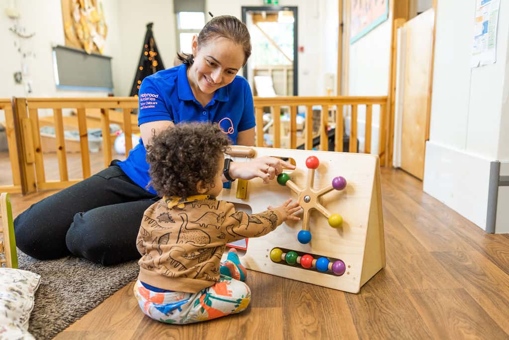 Bright smiling caregiver assisting toddler with educational wooden activity toy at Thrive Childcare, fostering early childhood development and learning in a safe, nurturing environment.