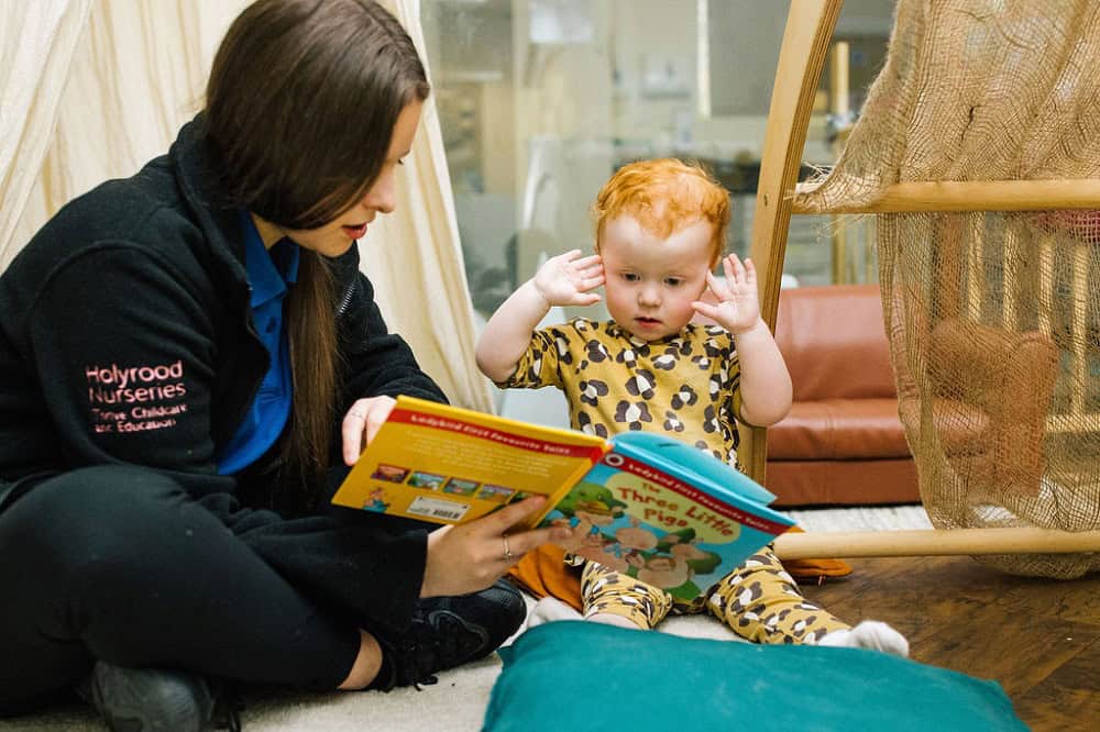 A young child with red hair and a caregiver reading a colourful children's book in a childcare setting.