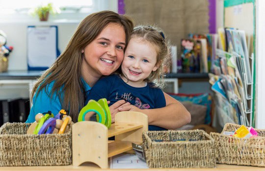 Bright smiling female caregiver with a young girl at Thrive Childcare, engaging in educational activities in a vibrant, colourful preschool environment.