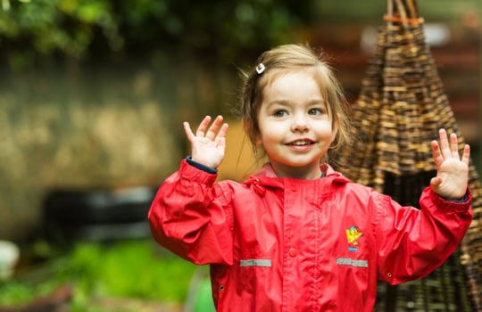 Young girl smiling outdoors at Thrive Childcare, wearing a red raincoat, engaging children-focused activities, promoting early childhood education and caring childcare services.