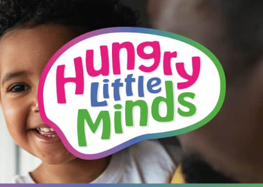 Bright smiling child with a balloon-shaped sign reading "Hungry Little Minds" at Thrive Childcare, promoting early childhood education and development in a welcoming nursery environment.