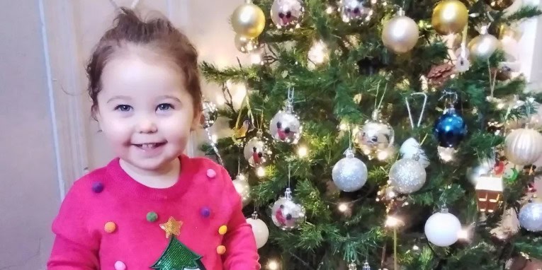 Bright smiling young girl in festive Christmas jumper next to decorated Christmas tree at Thrive Childcare, promoting holiday joy and early childhood celebration.