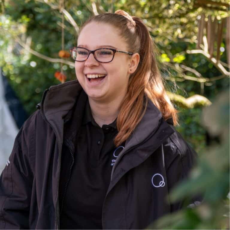 A smiling caregiver at Thrive Childcare outdoors, surrounded by greenery, representing nurturing early childhood education and caring childcare services.