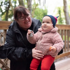 Bright smiling childcare worker with a young toddler outdoors at Thrive Childcare centre in a serene, nature-filled environment.