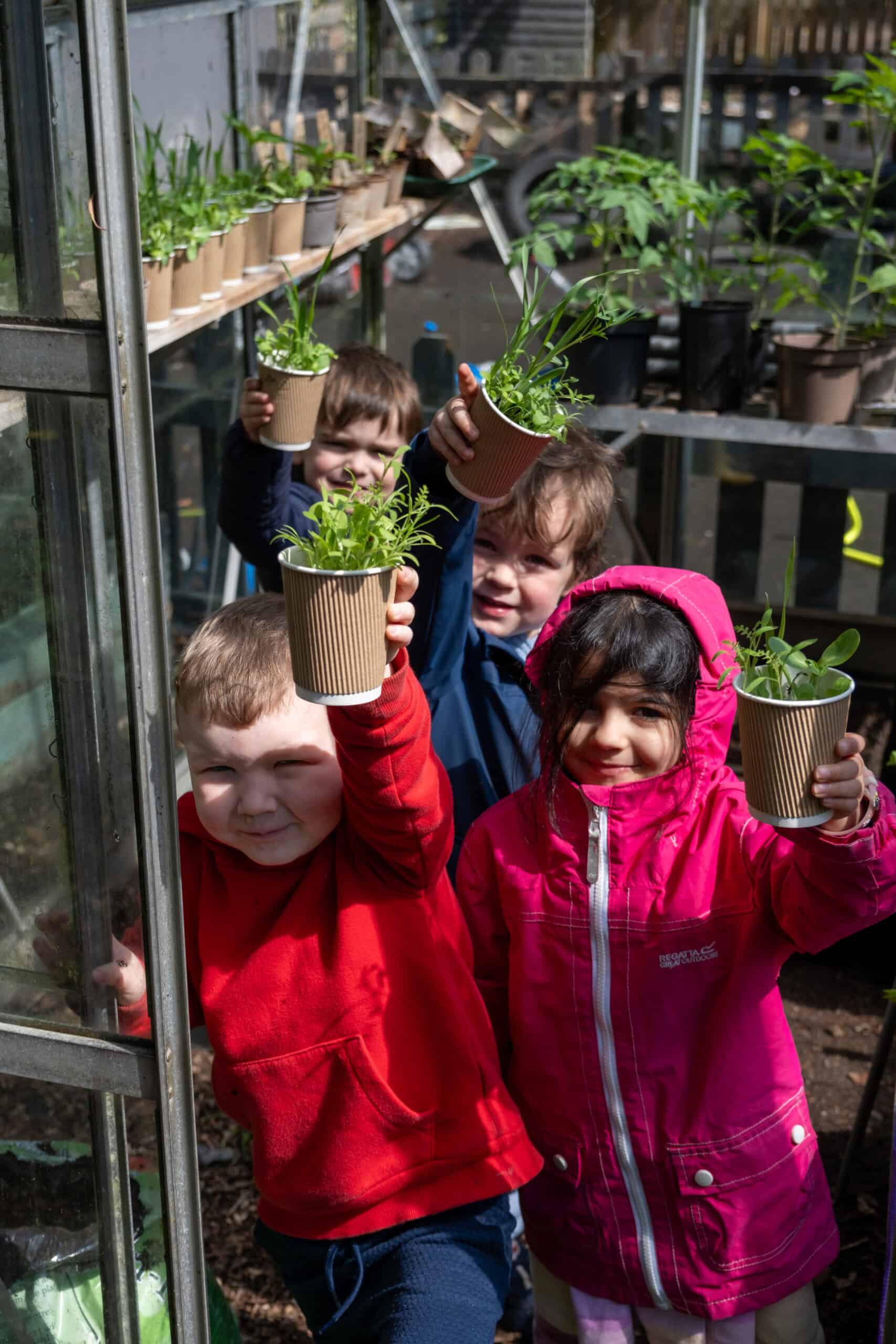 Bright children in colourful jackets holding potted plants, exploring nature at Thrive Childcare's outdoor garden nursery, promoting early environmental education and outdoor learning for kids.