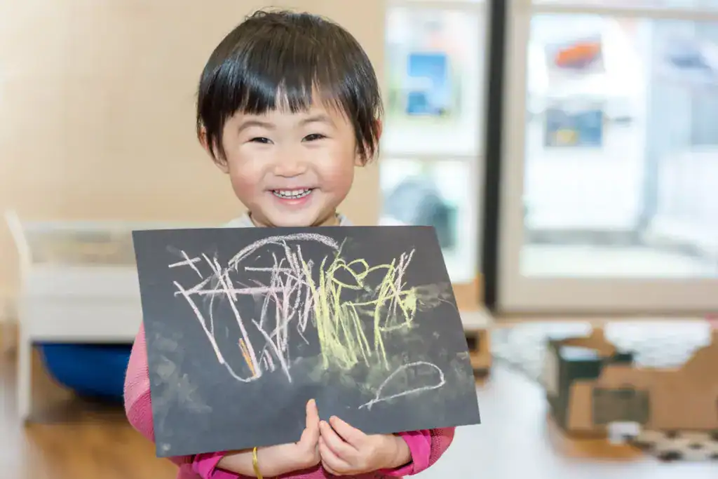 Smiling young child holding colourful chalk drawing on black paper in childcare setting, demonstrating early childhood education and creative play at Thrive Childcare.