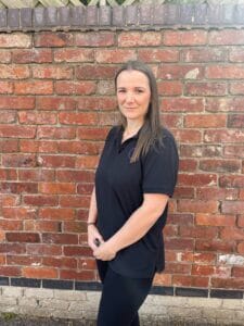 A confident childcare professional at Thrive Childcare standing outdoors against a brick wall, ensuring a safe and nurturing environment for children.