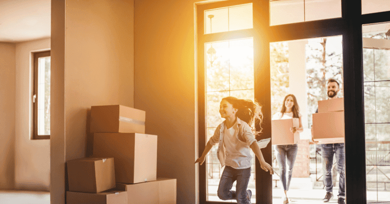 A woman and children entering a home with moving boxes, sunlight streaming through large windows.