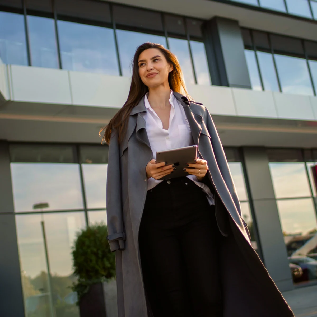 A woman in a long coat holding a tablet outside an office building