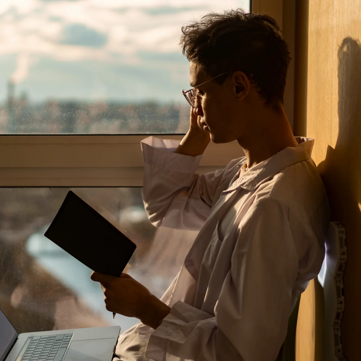 A man in a white shirt reading a document by a window, laptop beside him