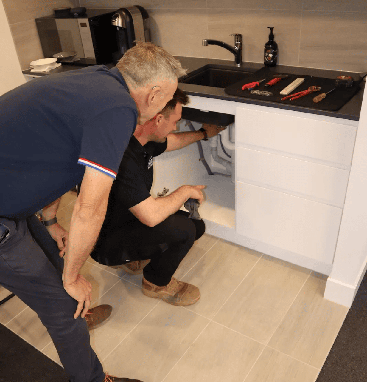 A plumber kneels under a kitchen sink, inspecting the pipes while another man observes.
