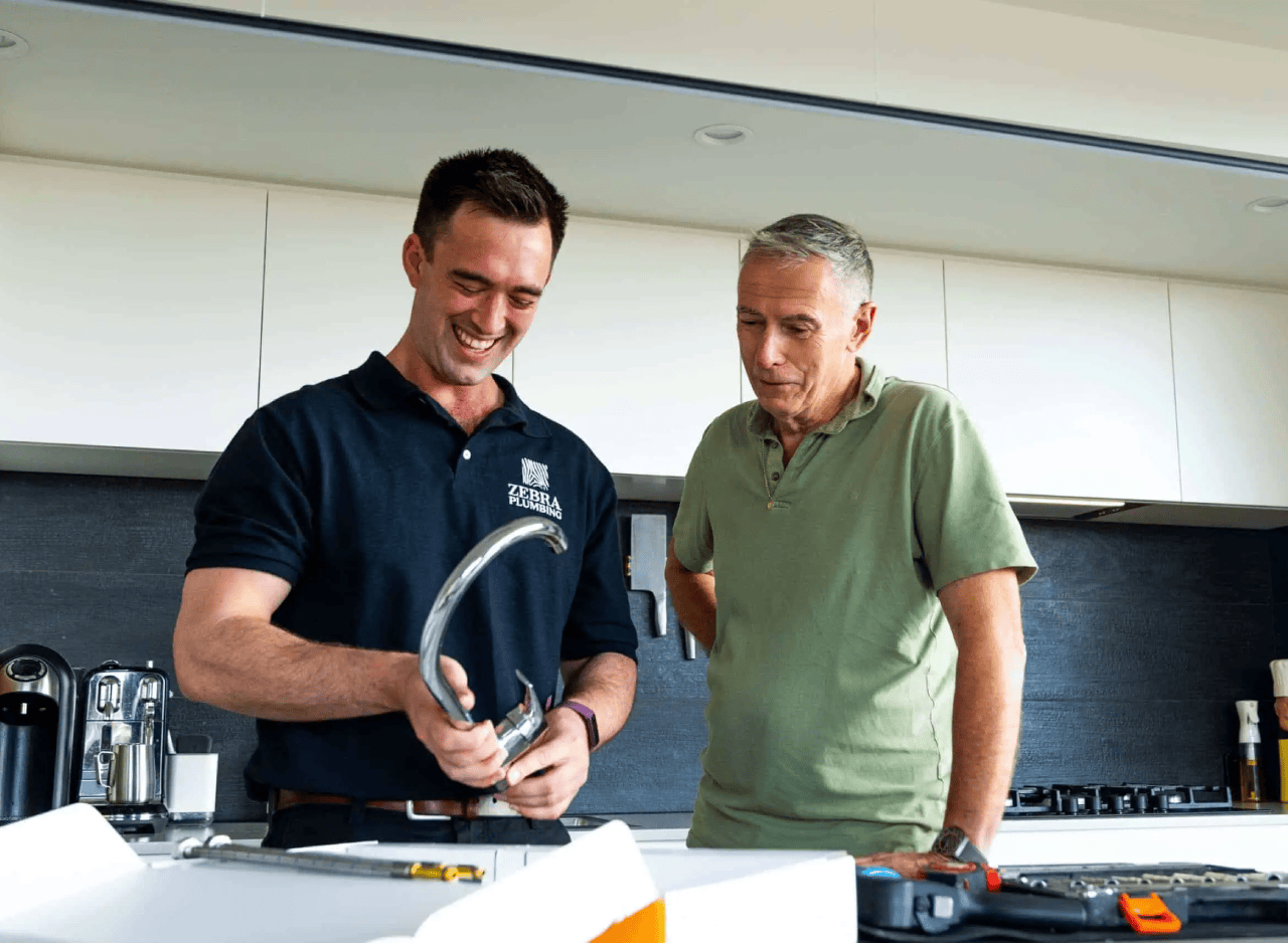 A smiling plumber in a Zebra Plumbing uniform holds a chrome kitchen tap while standing next to a customer in a green shirt.