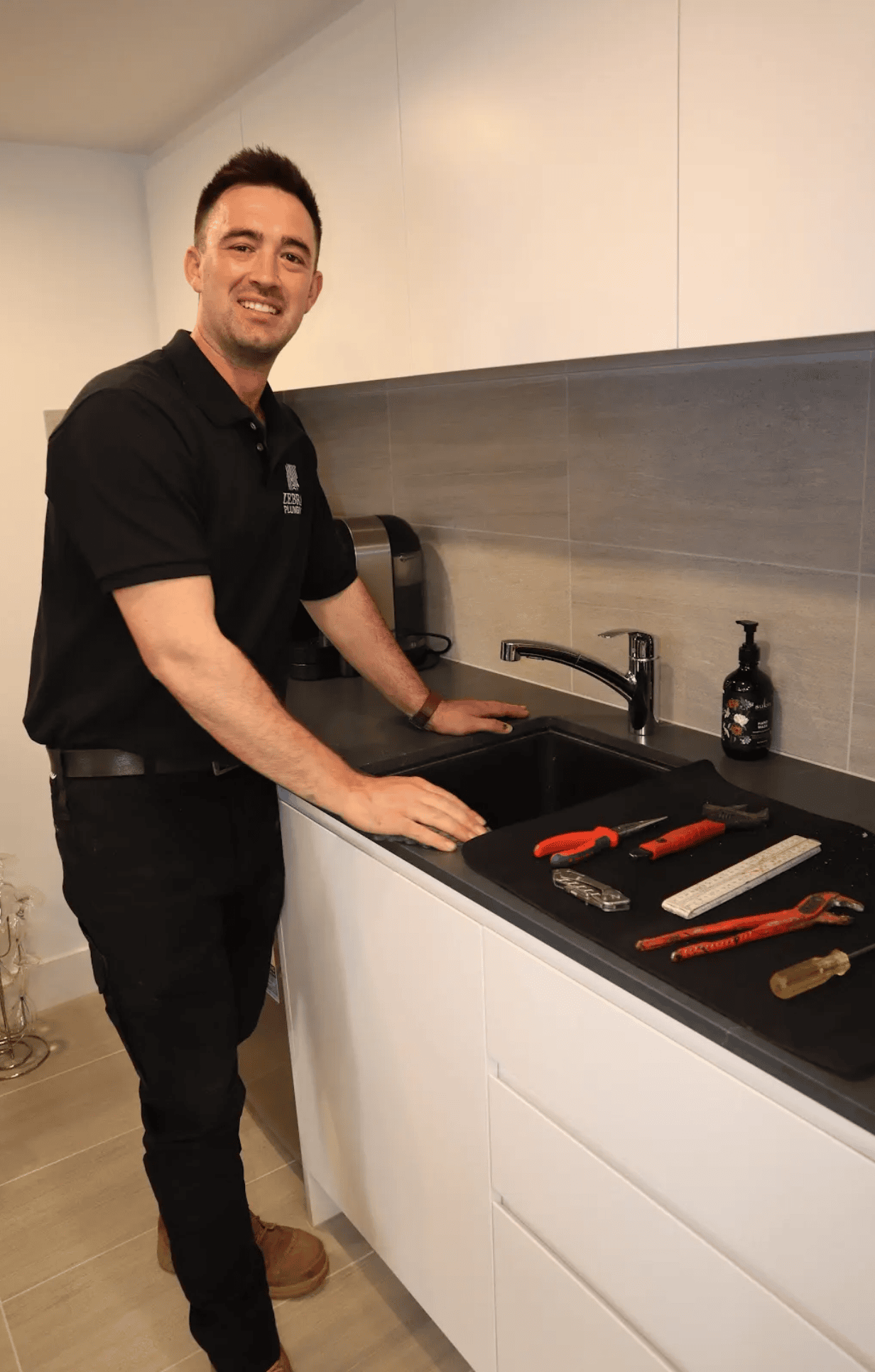 Smiling plumber beside a kitchen sink with plumbing tools neatly arranged on the benchtop.