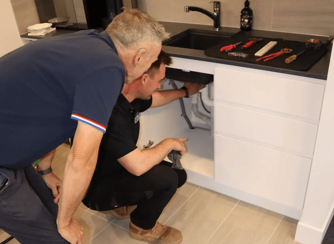 Zebra Plumbing technician inspecting pipework under a kitchen sink while explaining the issue to a customer.