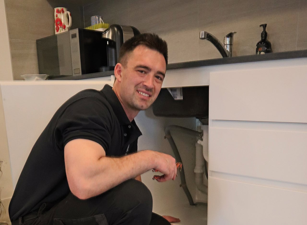 Plumber in blue uniform fixes a blocked drain under the kitchen sink.