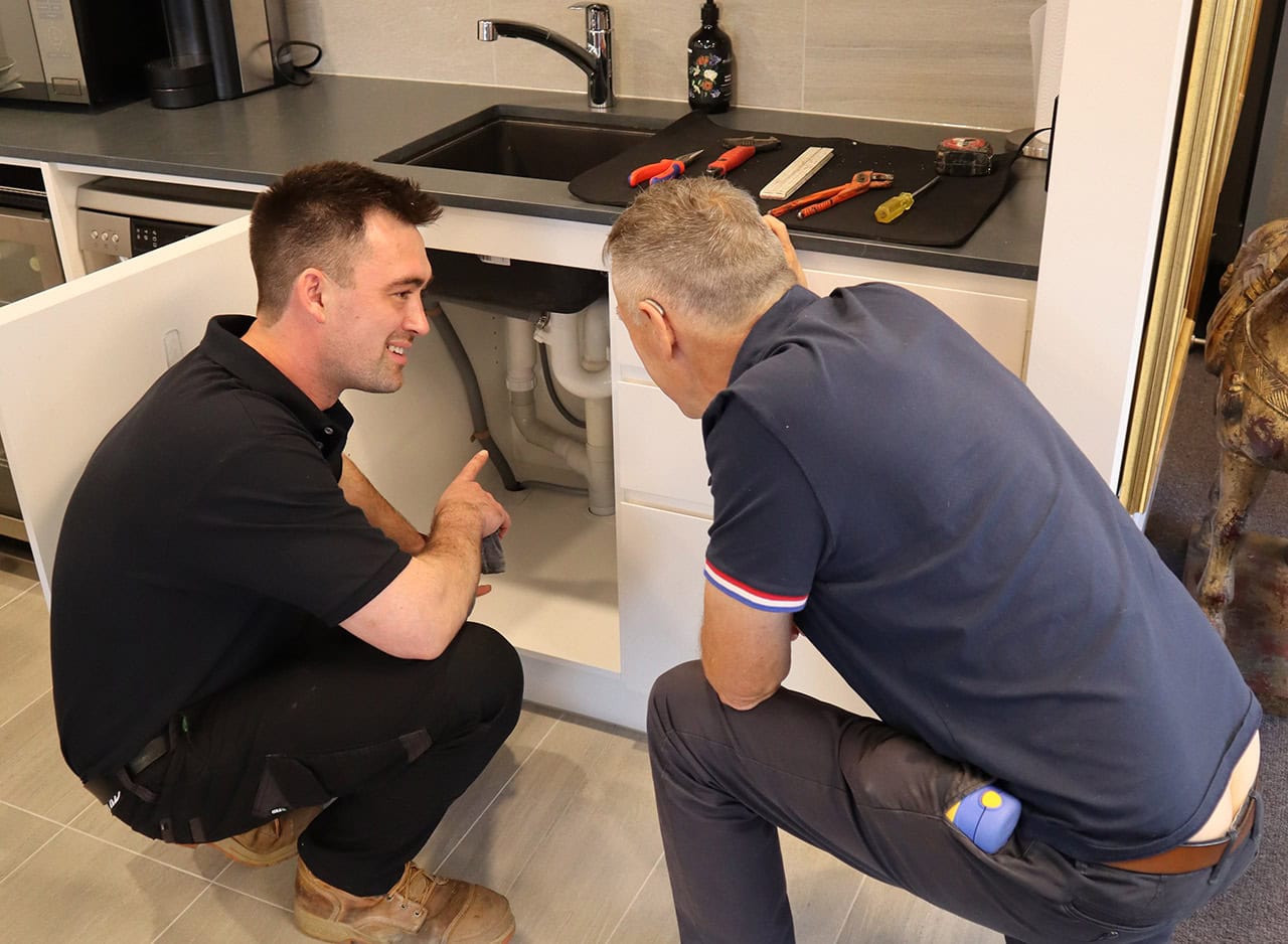 Plumber explains to a homeowner the pipes under the kitchen sink before working on the blocked drain.
