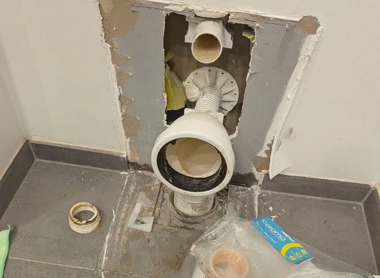 Toilet plumbing repair or installation with exposed pipes and wall opening.