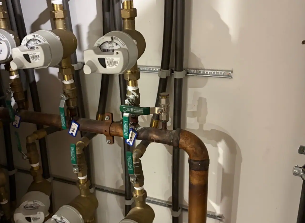 Residential water meter manifold with multiple brass pipes, white meter gauges, black insulation tubing, green valve tags,