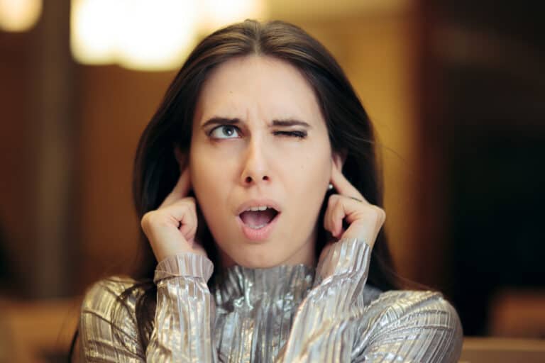 Woman closing her ears, with mouth open and one eye closed. Annoyed.
