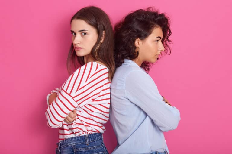 Picture of offended brunette girls, wearing casual outfits stand back to back with arms crossed and expressing argument, do not want to talk, isolated over pink background. People relationship concept