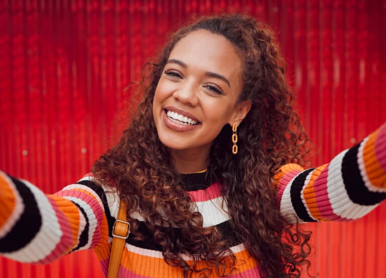 Face, selfie and smile with a black woman happy against a red background while taking a photograph. Social media, fashion and beauty with an attractive young female posing for a picture outdoor.