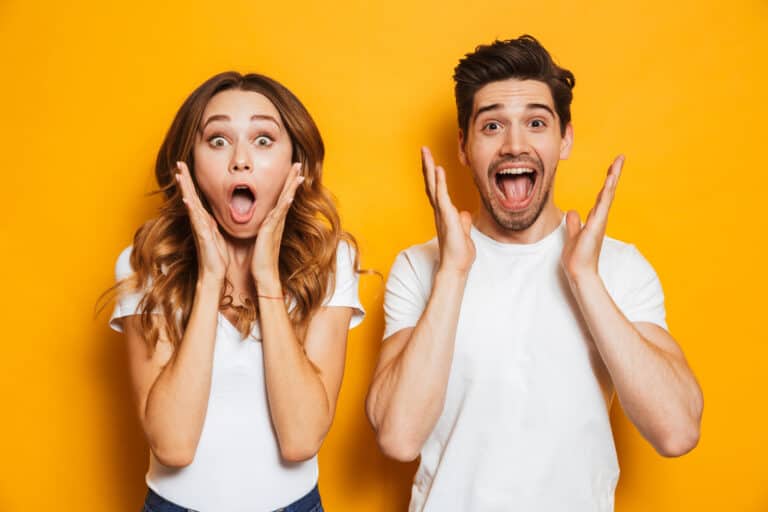 Photo of excited couple man and woman in basic clothing shouting in surprise or delight and raising arms isolated over yellow background