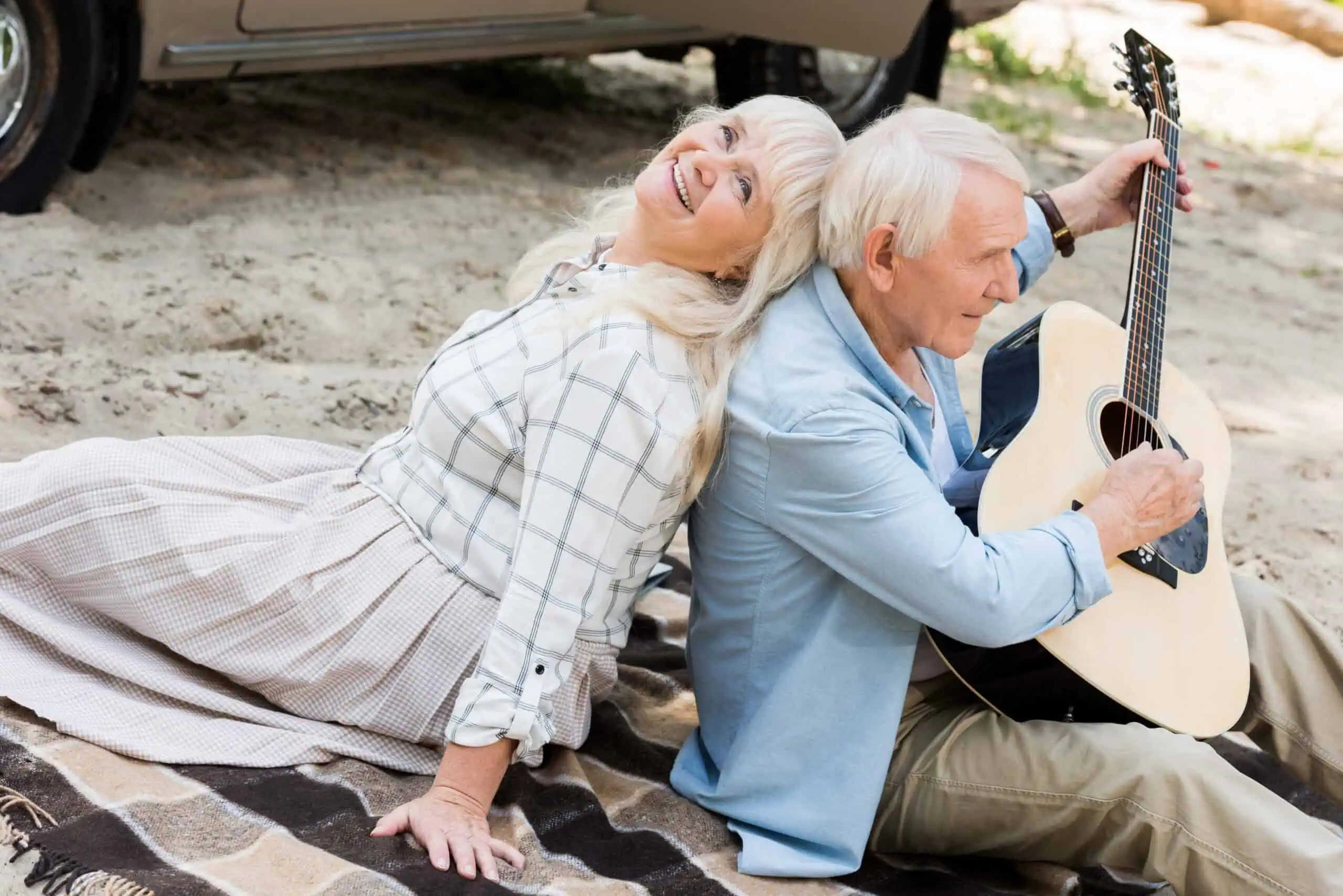 Senior woman sitting back to back with man playing guitar on sand
