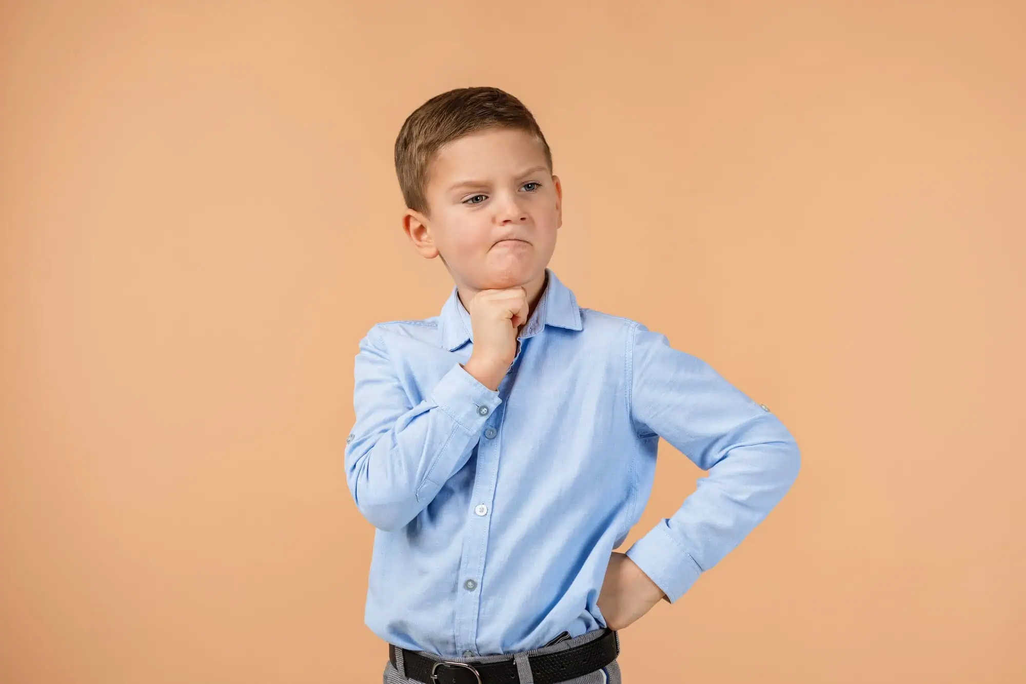 Cute little child boy in blue shirt thinks about something on beige background. Human emotions and facial expression