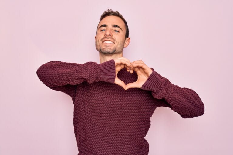 I handsome man with blue eyes wearing casual sweater standing over pink background smiling in love doing heart symbol shape with hands. Romantic concept.