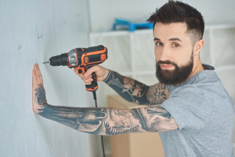 Tattooed man doing hole in wall with screw gun in new apartment, inexperienced millennial concept