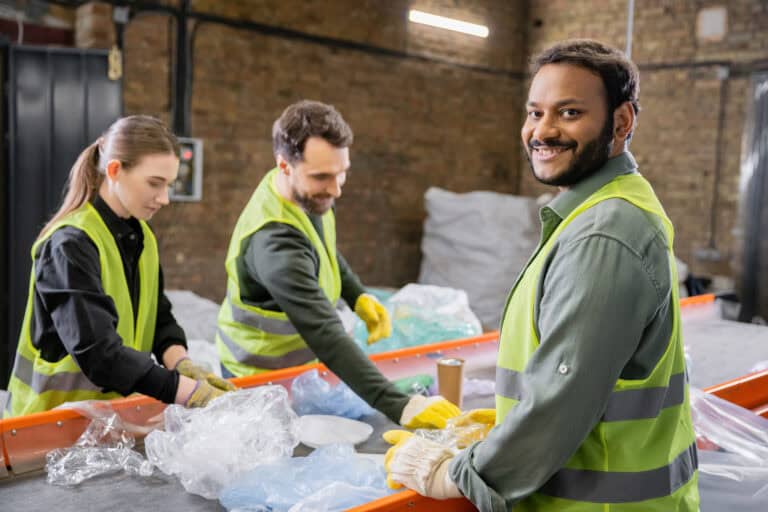Joyful indian worker in protective vest and gloves looking at camera while standing near trash on conveyor
