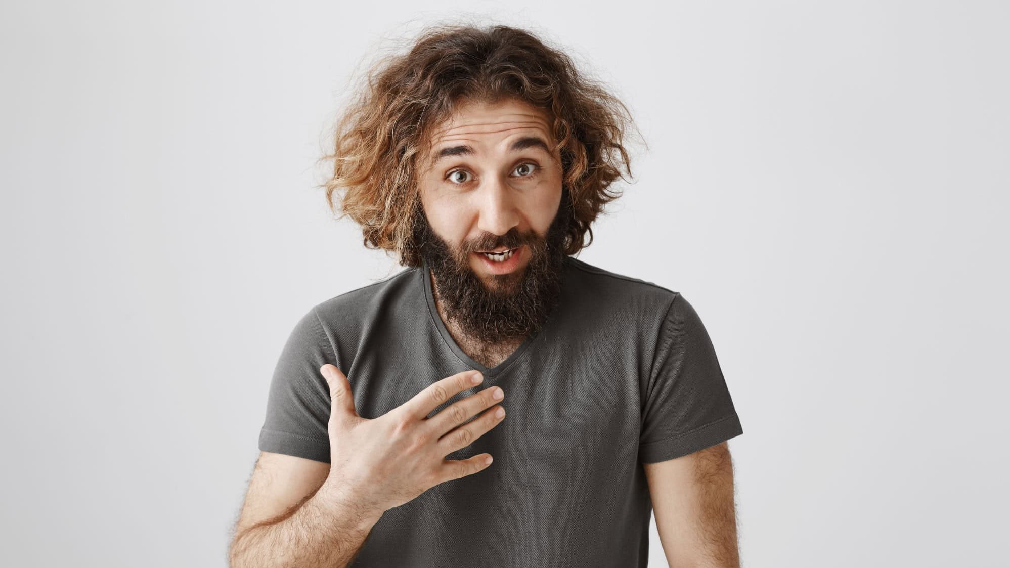 So it is my fault. Studio shot of displeased offended curly-haired male with beard pointing at chest and looking from under forehead with shocked and disbelieving expression, standing over gray wall